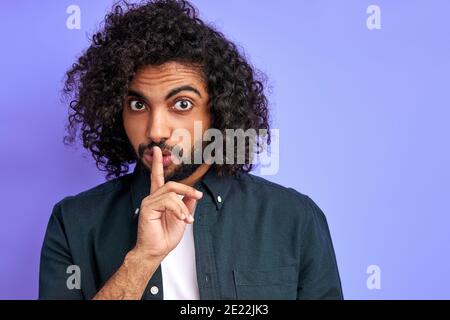curly arabic guy keep silence, show silence gesture, hold finger near mouth, look at camera, warning Stock Photo