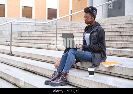 University female african student wearing protective face mask studying on her laptop sitting on stairs outside on campus. New normal in college. Stock Photo