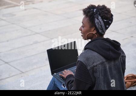 University female african student smiling and writing on her laptop sitting on stairs backwards outside on campus. College life concept. Stock Photo