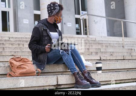 University female african student wearing protective face mask studying sitting on stairs outside on campus. New normal in college. Stock Photo