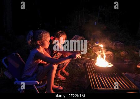 two girls roasting marshmallows over open fire, camping in summertime Stock Photo