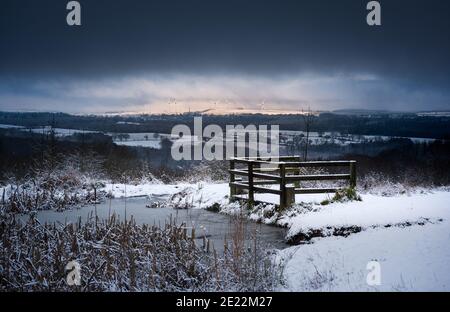 Winter snow covered landscape with frozen pond dark ominous cloudy sky and distant sunlit uplands with far off wind turbines in the sunlight Stock Photo
