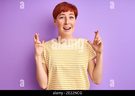 redhead adult woman hope for the best, keeping fingers together, good luck. isolated over purple background Stock Photo