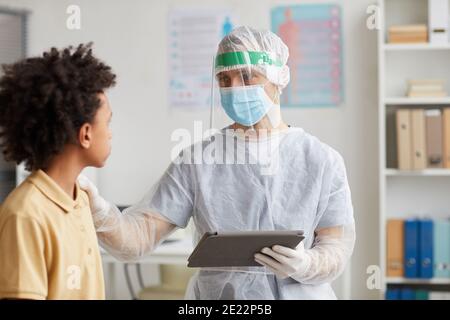 Portrait of doctor wearing full protective gear talking to African-American boy during consultation in clinic, copy space Stock Photo