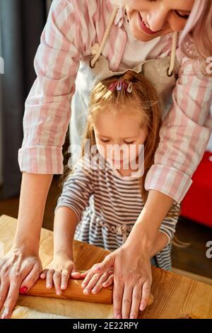 mom showing daughter how to roll out dough for pie or cookies, little cute kid child learning muffins bans cooking process with happy mom in light kit Stock Photo