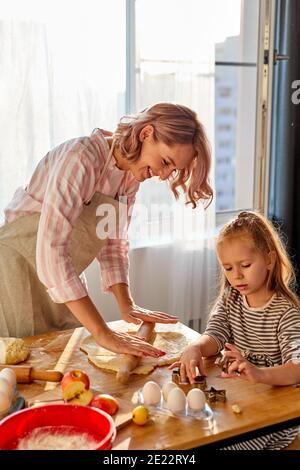 mother is teaching daughter to roll out dough for baking cookies, in the kitchen at home. on table Stock Photo