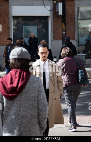 Young man crossing the street dressed in a raincoat, white shirt and black tie while other people are out of focus. Lifestyle concept. Stock Photo