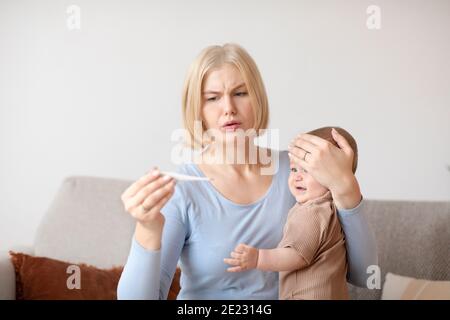 Anxious mother checking temperature of sick crying baby at home Stock Photo