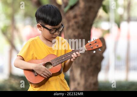 An Asian little boy wearing glasses is happily playing the ukulele. Asian little child is trying to play the ukulele with a fully happy moment. Stock Photo
