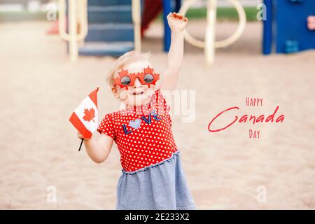 Happy Canada Day. Holiday card with greeting text. Adorable cute baby toddler girl holding Canadian flag. Child wearing funny maple leaves sunglasses Stock Photo