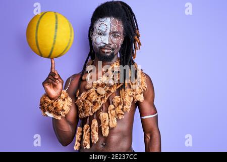 black man in national authentic wear holding yellow ball in hands, going to play basketball, sport game, having paintings on face. isolated on purple Stock Photo