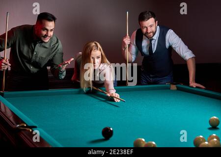 woman with two male friends are playing billiards in bar after work, have rest and leaisure time, preparing aiming to shoot pool balls Stock Photo