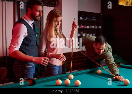young active friends are playing billiards in bar after work, have rest and leaisure time, preparing aiming to shoot pool balls Stock Photo