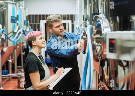Young workers of beer production factory standing in front of camera while woman making notes and looking at processing machine Stock Photo