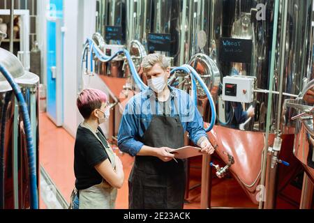 Two young brewers of large contemporary beer production factory consulting about some working moments between steel tanks Stock Photo