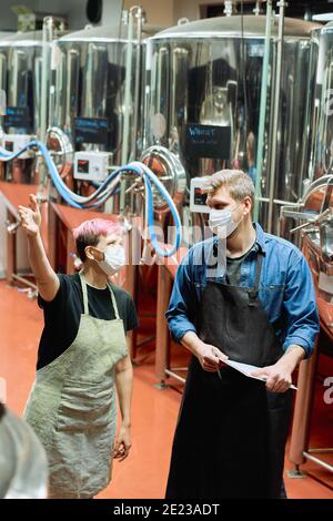 Two young brewers in protective masks consulting about some working moments while female worker pointing at one of large steel tanks Stock Photo