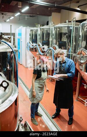 Young brewery master pointing at document while explaining his female subordinate which ingredients she should use for beer production Stock Photo