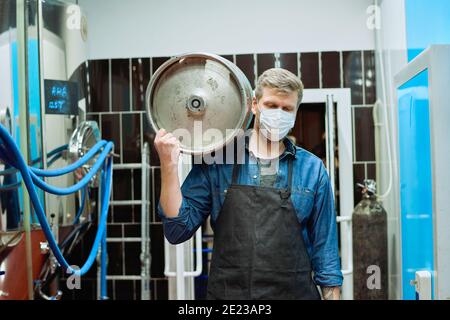 Young male worker of beer production plant in workwear and protective mask carrying cistern on shoulder while moving along large steel tanks Stock Photo