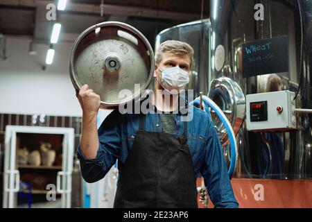 Male worker of contemporary beer production plant in workwear and protective mask carrying cistern while moving along large tanks Stock Photo