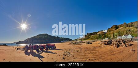 Panoramic image of La Greve de Lecq at low tide with a clear sky early morning with the sun shining with tractors on the beach, sandy beach and rocks. Stock Photo