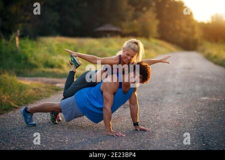 Training together is more fun. Young handsome man doing push ups while his girlfriend is sitting on his back. Stock Photo