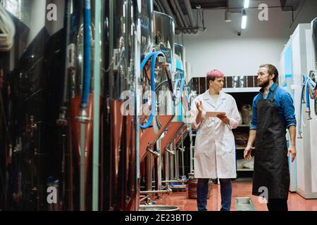 Young female brewery expert in whitecoat using digital tablet during discussion of characteristics of new equipment with male brewer Stock Photo
