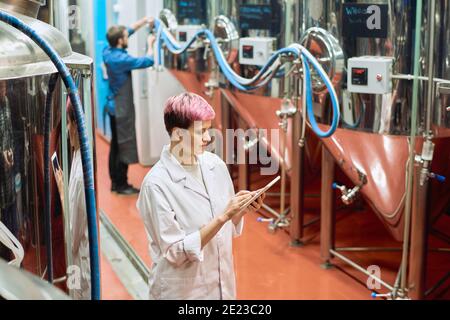 Young female brewery expert looking through data in digital tablet while standing by new equipment for beer production against male worker Stock Photo