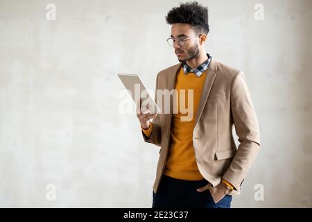 Young mixed-race elegant man in smart casualwear standing in front of camera and looking at display of digital tablet Stock Photo