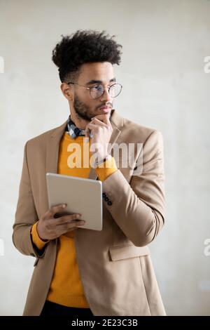 Young mixed-race pensive businessman in smart casualwear using digital tablet in front of camera against grey background Stock Photo