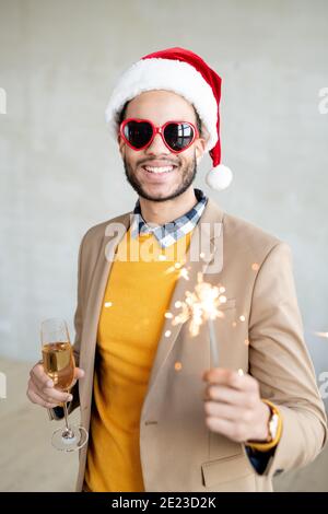 Happy young businessman in casualwear, Santa cap and heartshaped sunglasses holding flute of sparkling champagne and bengal light Stock Photo