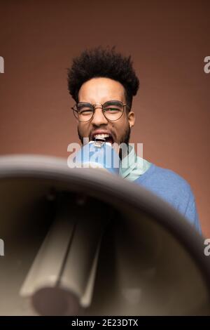 Young furious mixed-race businessman or boss in eyeglasses shouting loudly in megaphone at someone over brown background Stock Photo