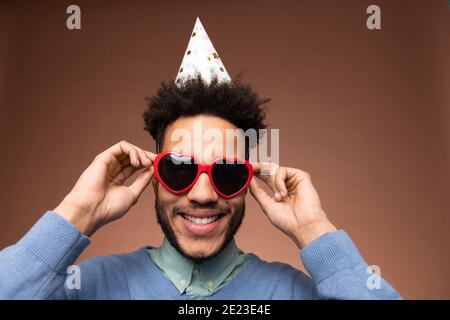 Young smiling mixed-race man in smart casualwear and birthday cap touching heartshaped sunglasses while standing in front of camera Stock Photo