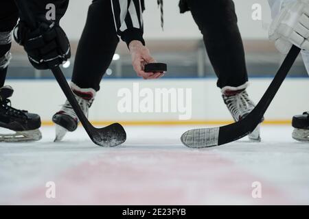 Hand of referee holding puck over ice rink with two players with sticks standing around and waiting for moment to shoot it Stock Photo