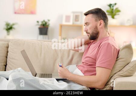 Side view of young sick man with thermometer sitting on couch under blanket, touching his neck and consulting online doctor in front of laptop Stock Photo