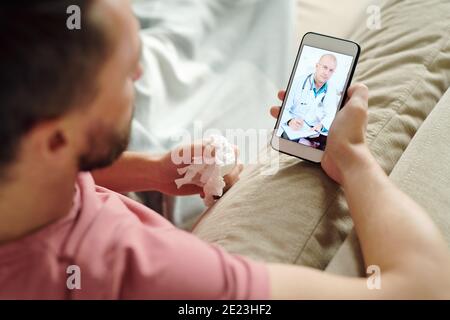 Confident mature male doctor on smartphone screen making notes and giving medical recommendations to young sick man lying on couch Stock Photo
