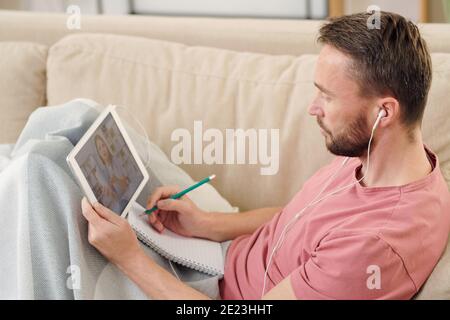 Young sick man in earphones sitting on couch under blanket, making notes and consulting online doctor while holding digital tablet on his knees Stock Photo