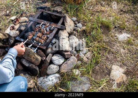 Delicious appetizing grilled meat, cooked over a fire in the courtyard of a country house. Male hands take skewers and turn them for better cooking. Stock Photo