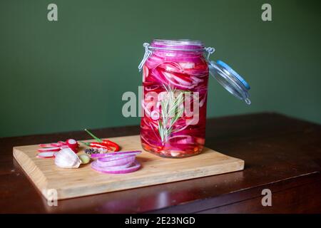 Homemade sweet and spicy pickled red onions sliced in a large glass jar, with the ingredients beside on a wooden cutting board with chili peppers, gar Stock Photo