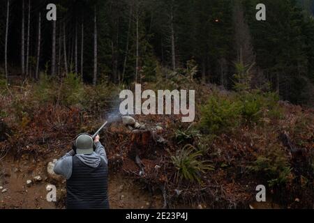 A mean wearing a vest, hat and ear protection holds an old 12 gauge shotgun, aiming and shooting at orange clay pigeons to practice. Pump action shotg Stock Photo