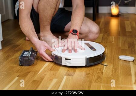 Caucasian young man doing white robot vacuum cleaner maintenance. Autonomous wireless modern vacuum cleaner. Male repairing robot cleaner home on floo Stock Photo