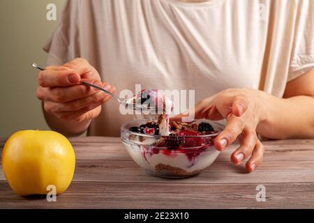 A caucasian woman is eating a fresh home made glass bowl of creamy yogurt parfait with berries, muesli and seeds in it on wooden table Close up photo Stock Photo