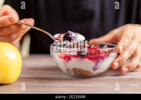 woman wearing black shirt eats a fresh homemade glass bowl of creamy yogurt parfait with berries, muesli and seeds in it on wooden table Closeup photo Stock Photo