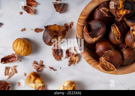 A wooden bowl of homemade oven roasted tsty chestnuts with shells and skins scattered on the marble table top. A traditional winter, autumn snack with Stock Photo