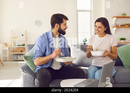 Married couple talking holding takeaway tray with food and having lunch at home sitting on sofa. Stock Photo