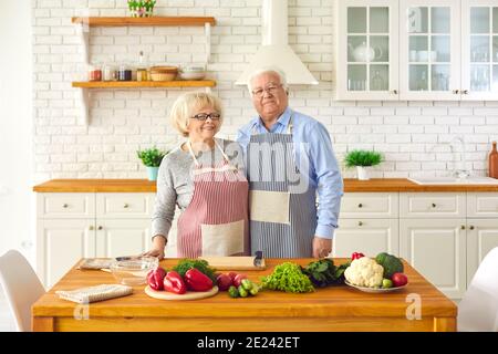 Senior couple standing in aprons in the kitchen near cooking products and looking at camera. Stock Photo