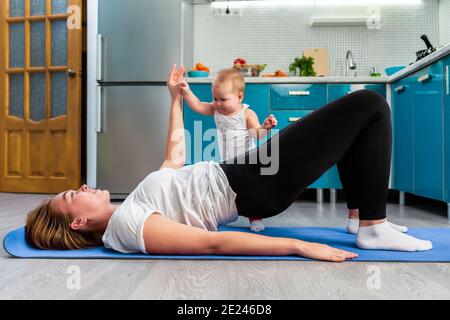 A mother with her baby is doing fitness on a mat in the kitchen. The child stands near the mother's leg and holds her hand. The concept of home sports Stock Photo