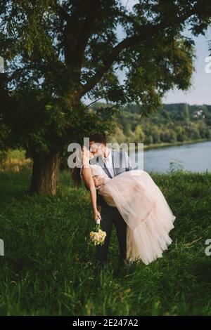 Beautiful in love just married couple kissing in nature. Bridegroom holding his bride in arms and kissing her. Standing in nature near river. Stock Photo
