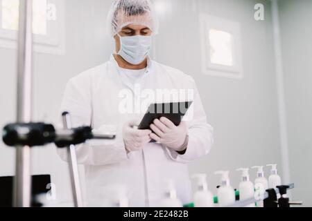 Picture of young man in sterile clothes using tablet . Controlling production line on factory. Bottles with cosmetic products on production line. Stock Photo