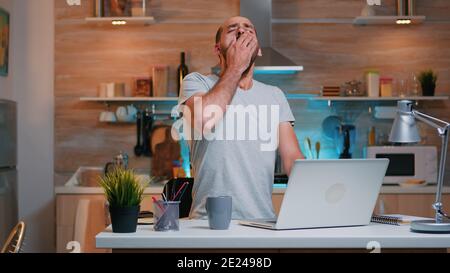 Tired sleepy man yawning, working at home desk in the kitchen. Busy overwork focused employee using modern technology network wireless doing overtime for job reading writing, searching Stock Photo