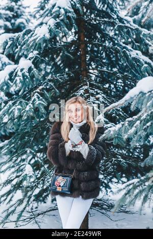 Smiling woman weared in fur coat walks in winter snowy park. Stock Photo